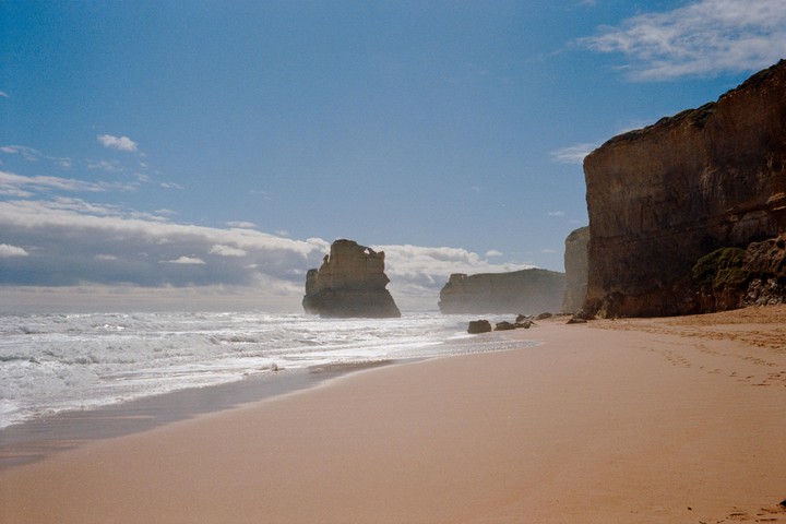 Waves crash on a sandy beach next to large rock formations and cliffs under a blue sky with clouds.