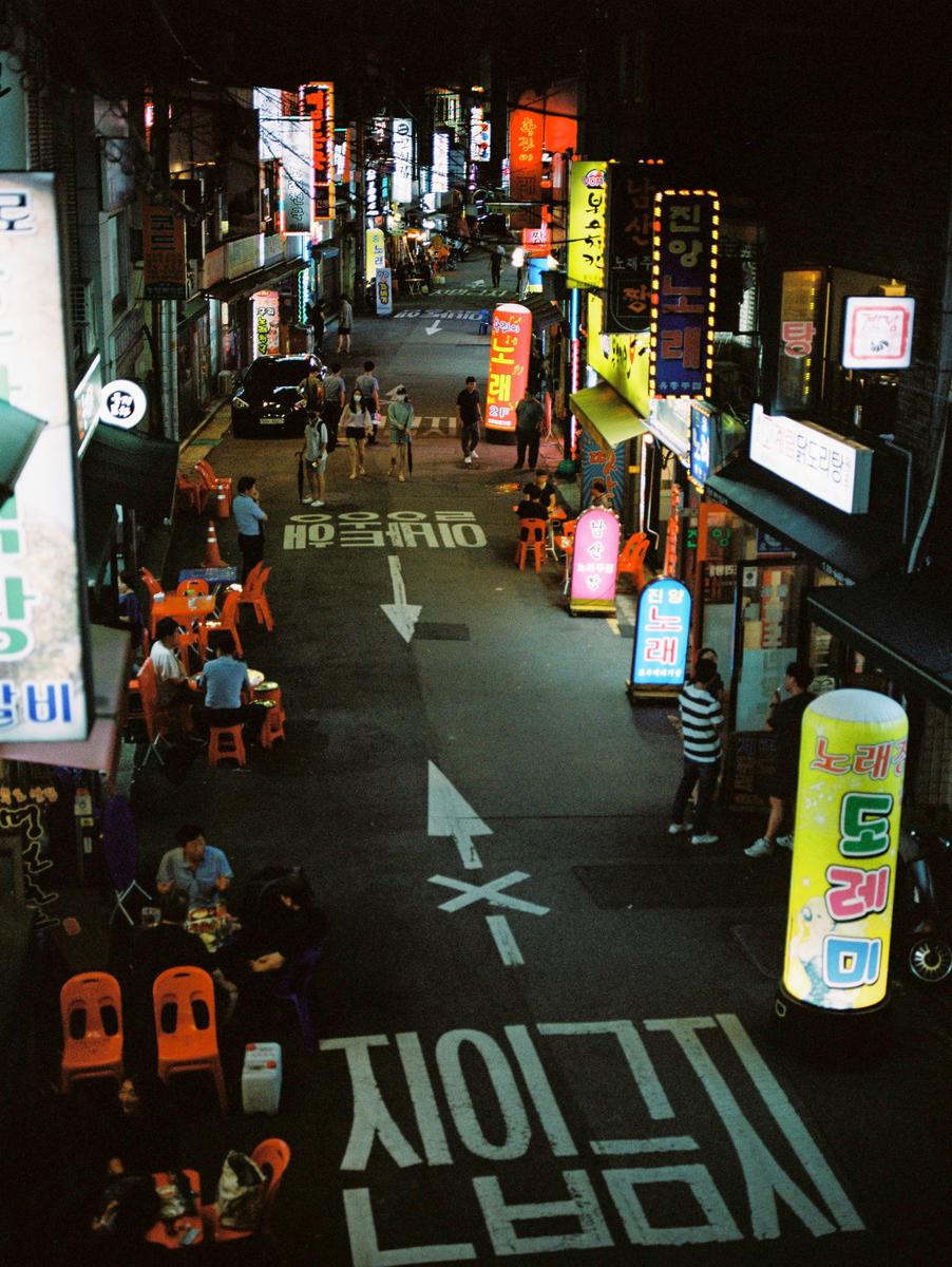 A street in Seoul is filled with people, neon signs, and outdoor dining at night.