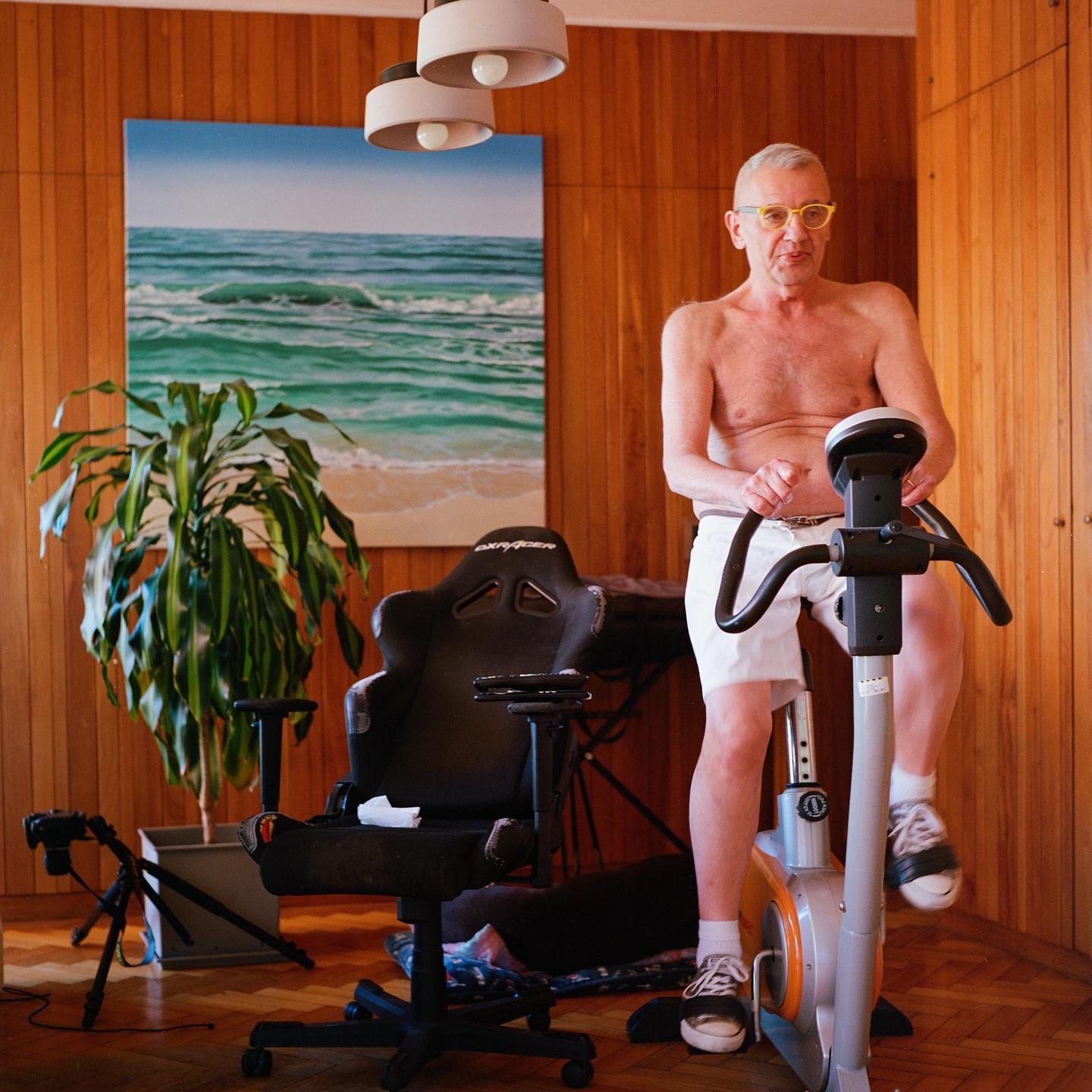 A man with yellow glasses is exercising on a stationary bicycle in a room with wood paneling and a painting of a beach.