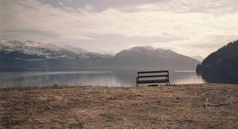A wooden bench sits on a grassy bank overlooking a lake with snow-capped mountains in the distance under a cloudy sky.