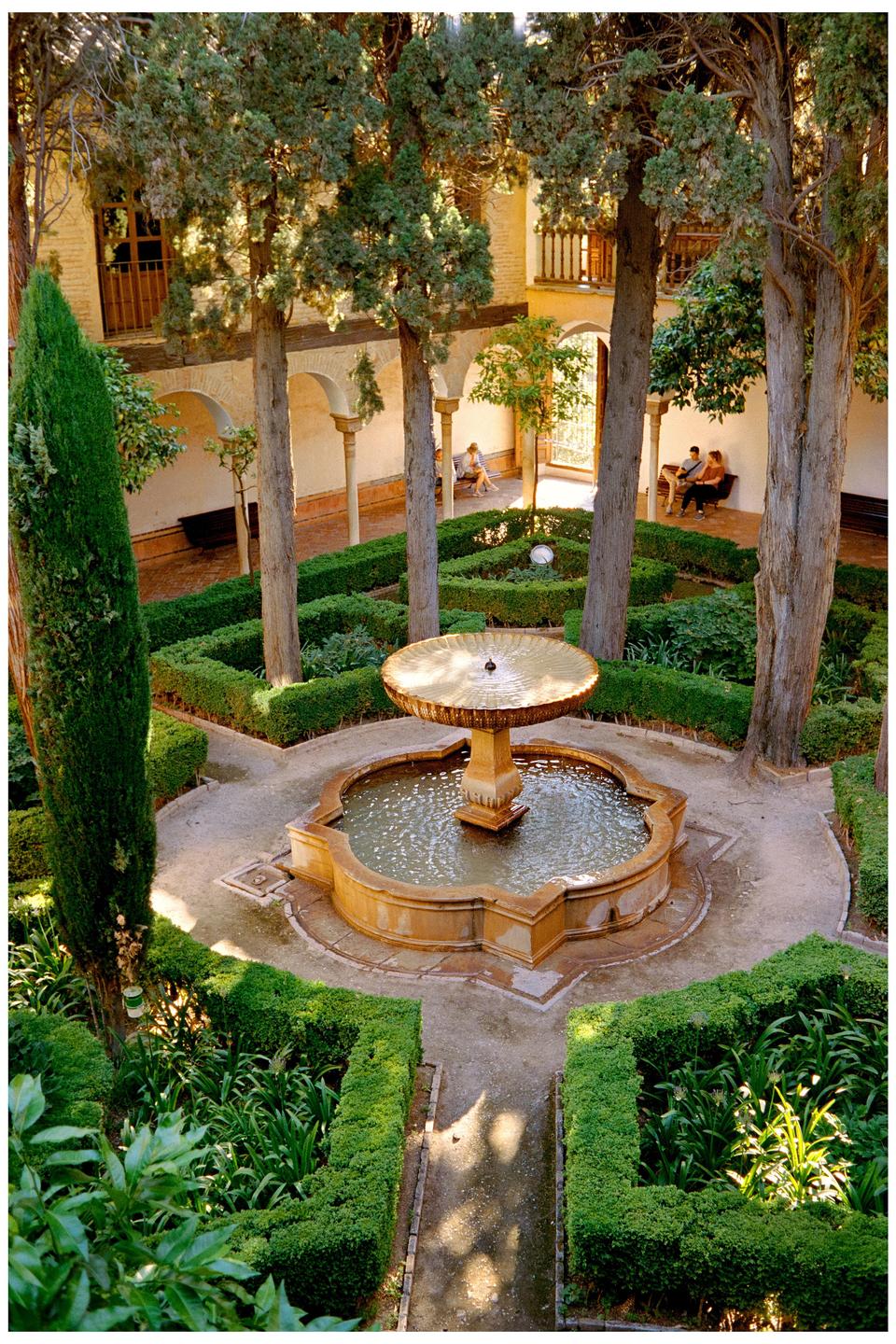 A courtyard garden with a fountain, trees, and manicured hedges is surrounded by a building with arches and balconies.