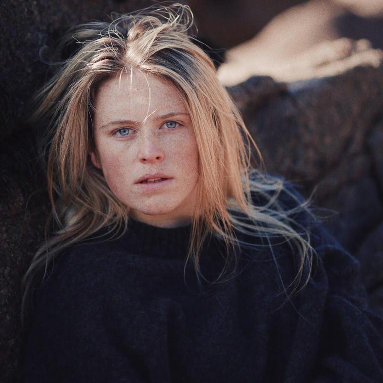 A woman with long blonde hair and freckles looks directly at the viewer.