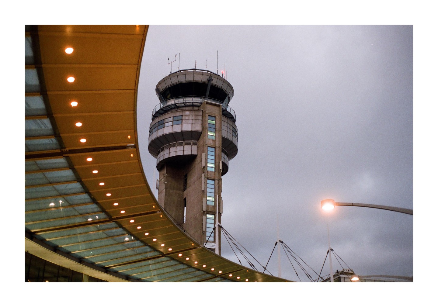 An airport control tower stands tall against a cloudy sky, with a curved, illuminated structure in the foreground.