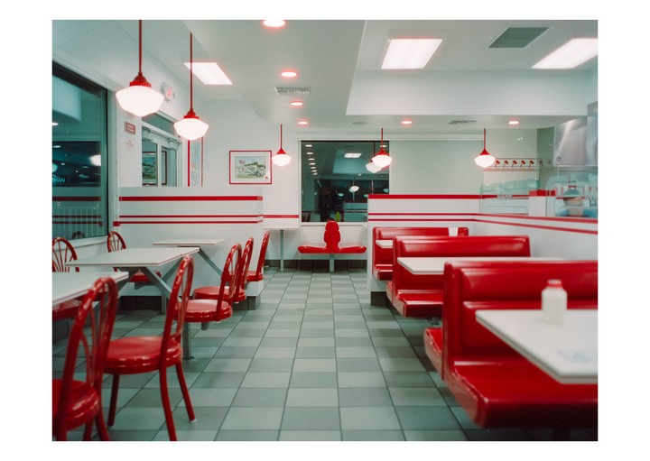 An empty diner with red booths and chairs is illuminated by overhead lights at night.