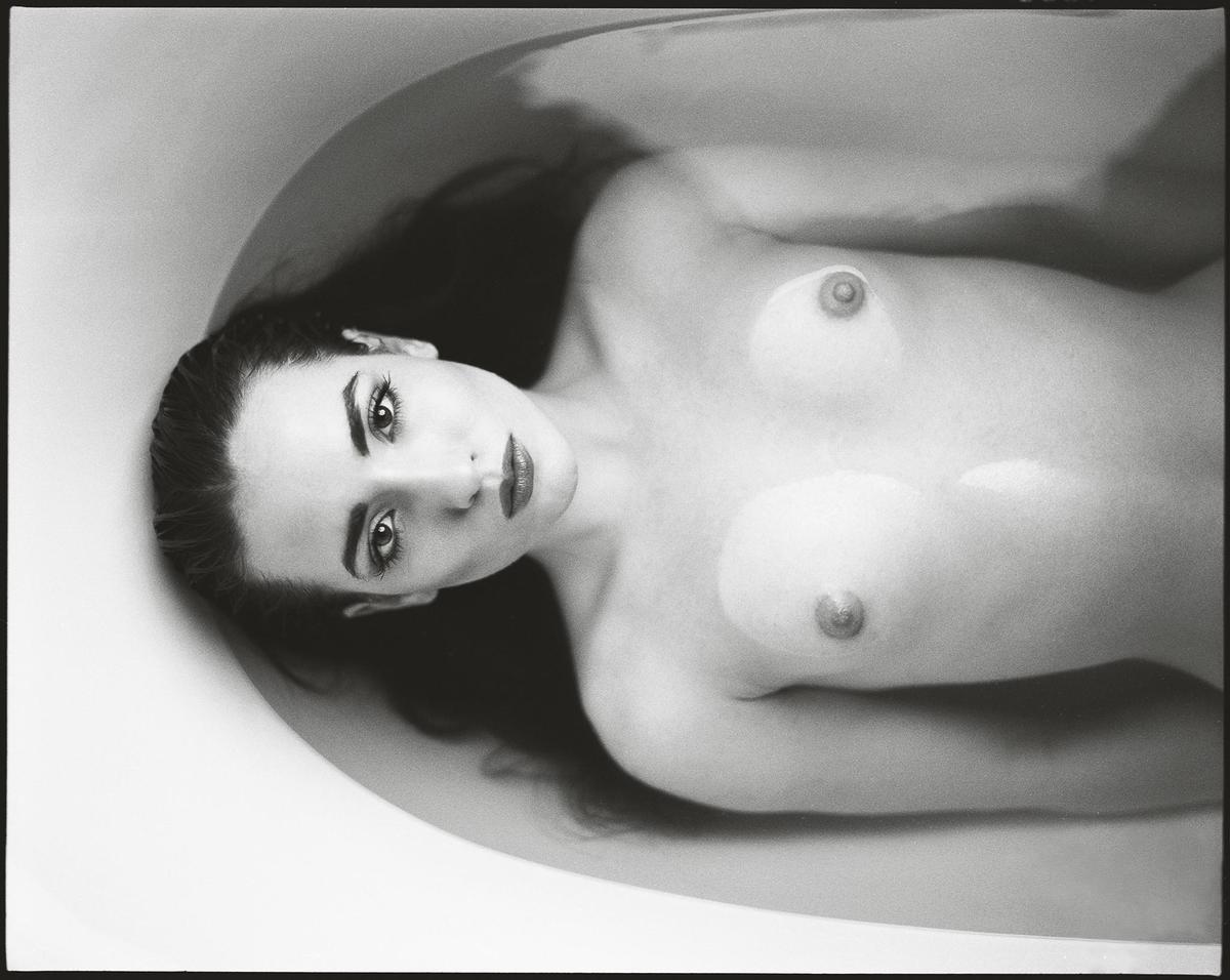 A woman with long hair is lying in a bathtub, looking up at the viewer.