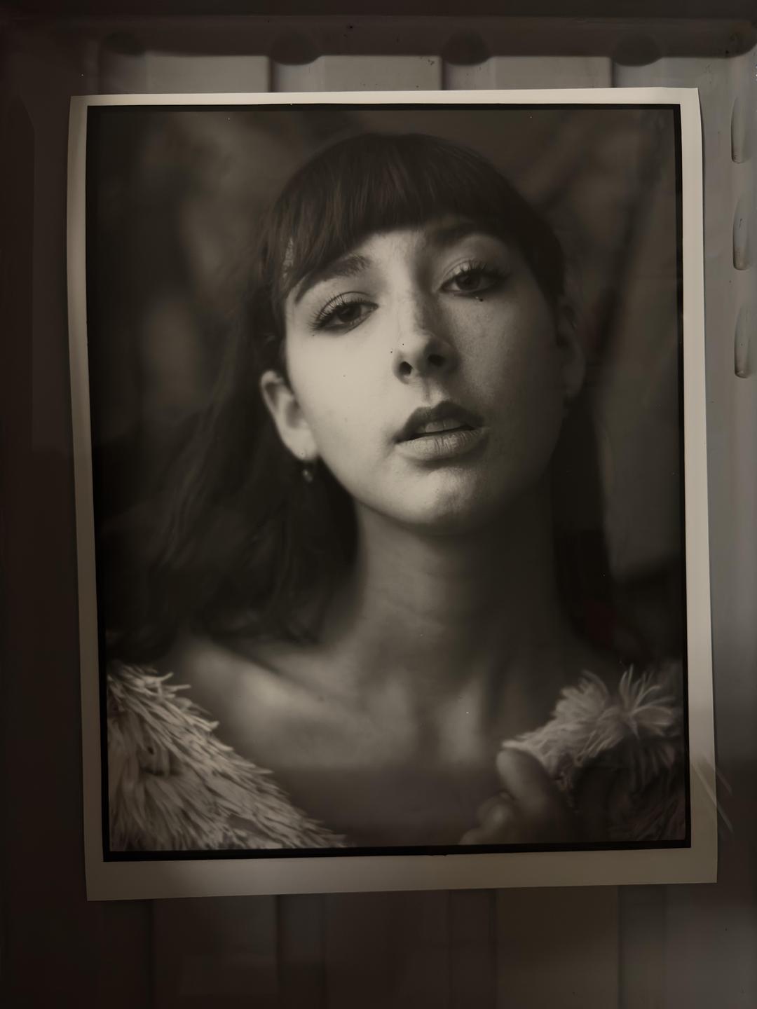 A woman with bangs and a fluffy garment looks directly at the viewer in a black and white portrait.