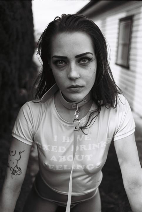 A woman wearing a collar and leash looks directly at the viewer.