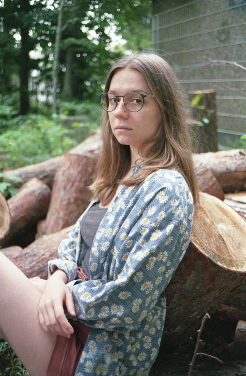 A woman wearing glasses and a daisy-patterned shirt sits among cut logs in a wooded area.