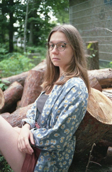 A woman wearing glasses and a daisy-patterned shirt sits among cut logs in a wooded area.
