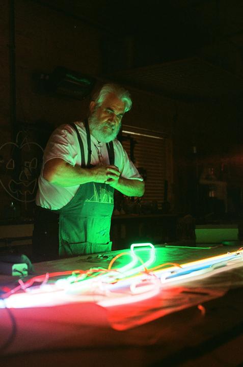 A man with a white beard and an apron stands over a table with neon lights.
