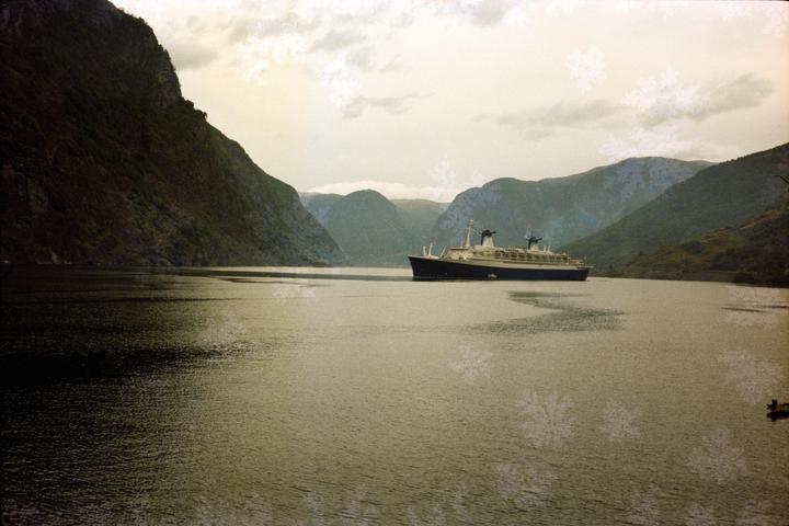 A large ship sails on a body of water surrounded by mountains under a cloudy sky.