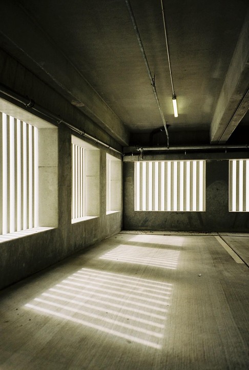 Sunlight streams through barred windows, casting striped shadows across the floor of a concrete parking garage.