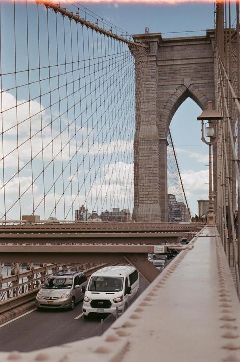 Cars drive on the Brooklyn Bridge with its cables and stone tower visible against a cloudy sky.