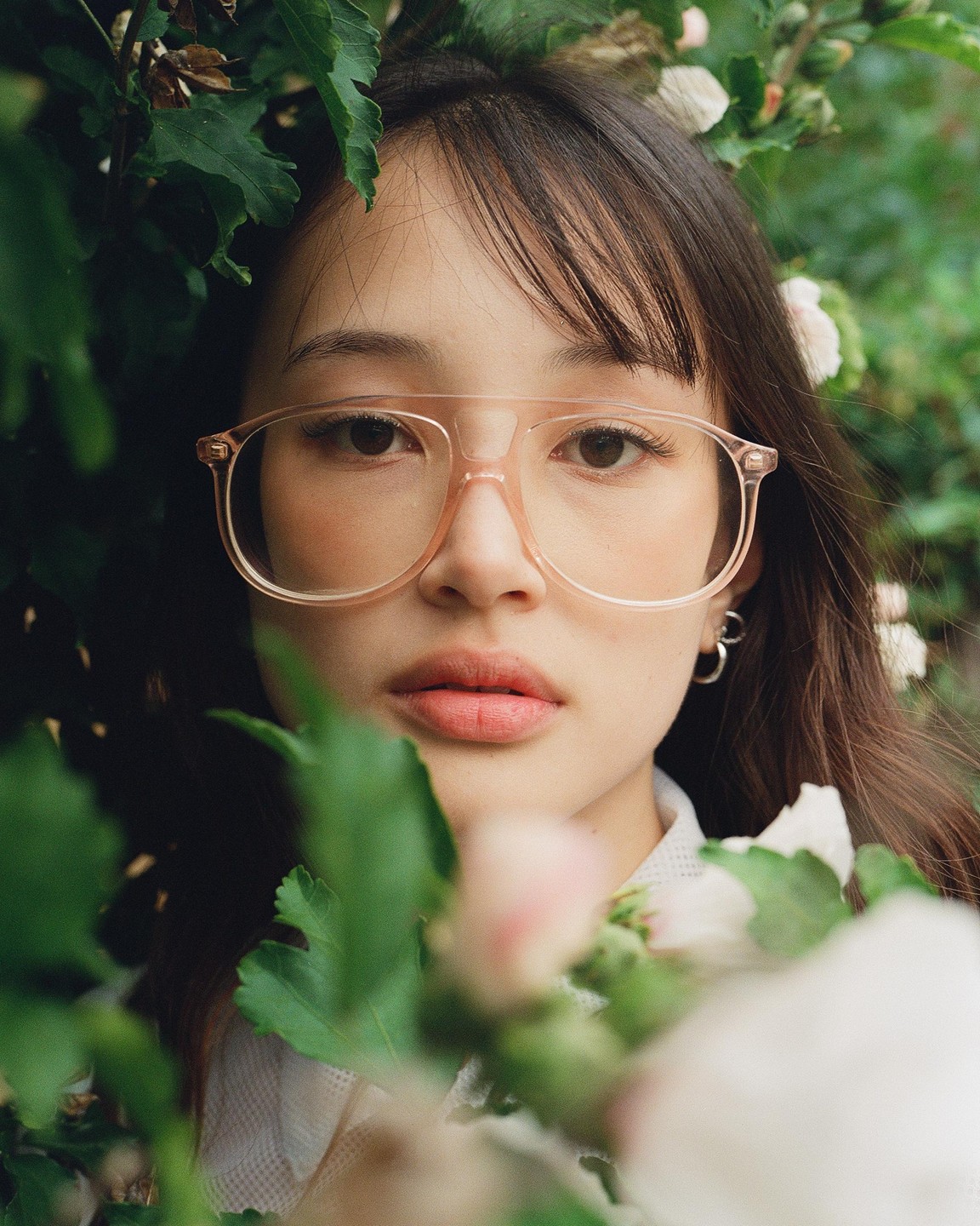 A woman wearing glasses looks directly at the viewer, framed by green leaves and white flowers.