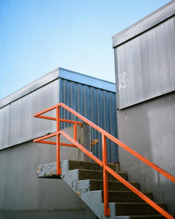 An orange handrail leads up a concrete staircase next to a corrugated metal building.