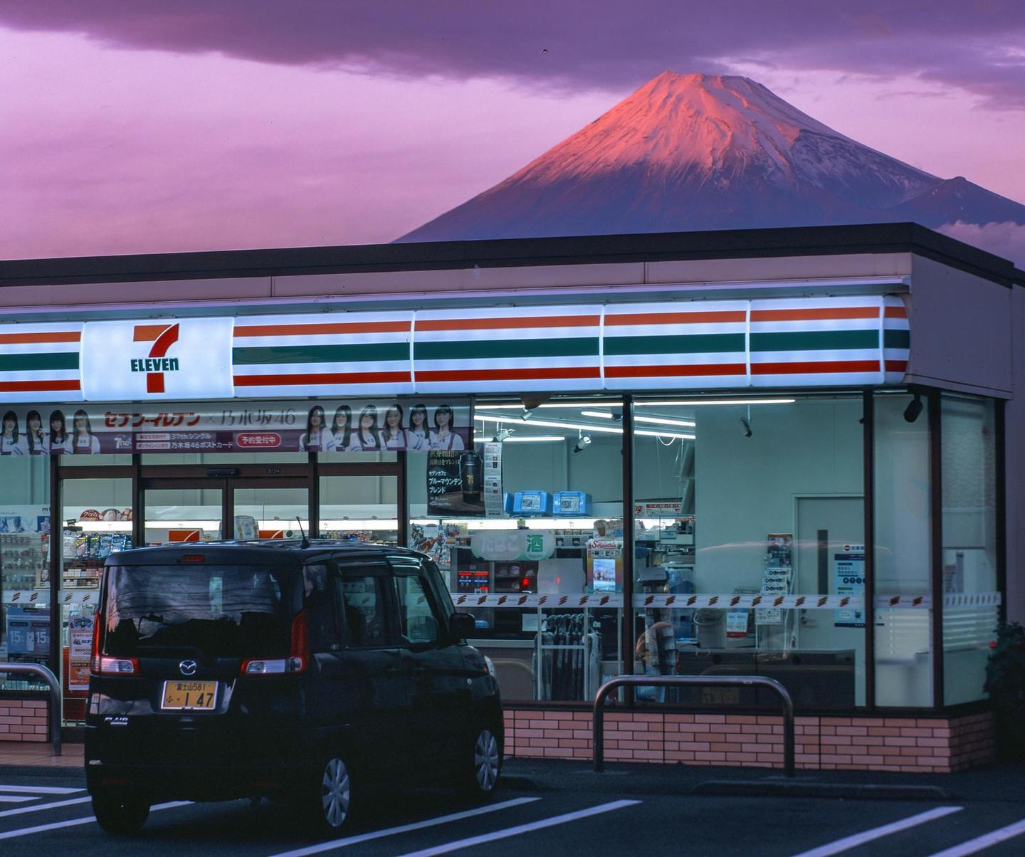 A black car is parked in front of a 7-Eleven convenience store with a mountain in the background under a purple sky.