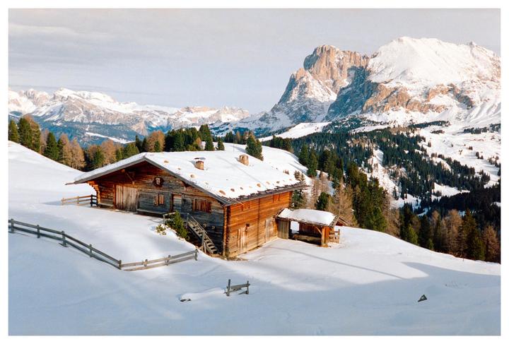 A wooden cabin with a snow-covered roof sits on a snowy hillside with mountains and trees in the background.