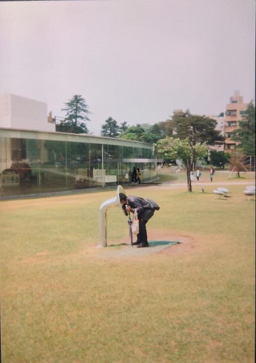 A man bends over a silver sculpture in a grassy park with a modern building in the background.