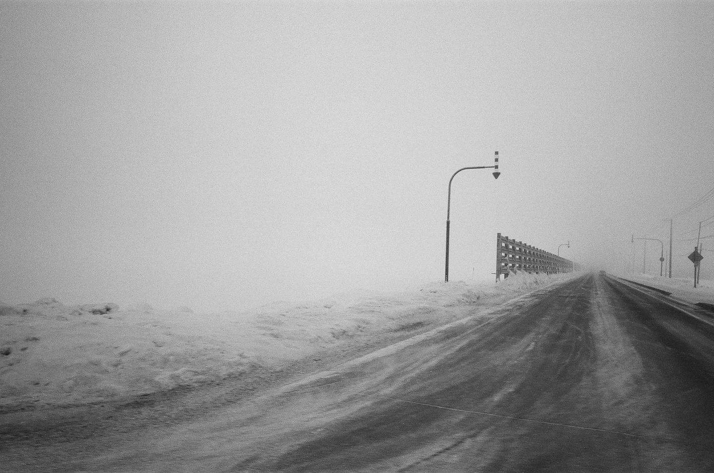 A snow-covered road stretches into the distance under a hazy sky, with a street light and a wooden fence visible on the right.