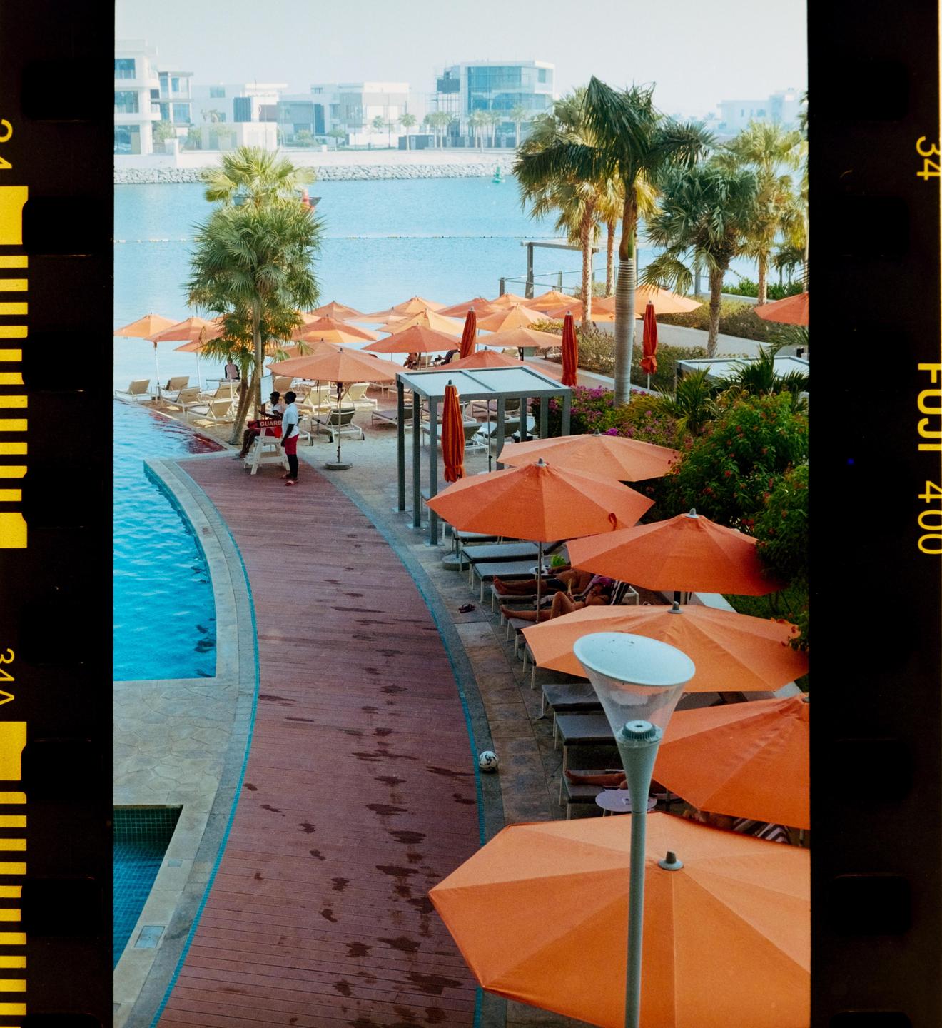 A resort with orange umbrellas and palm trees is next to a body of water with buildings in the distance.