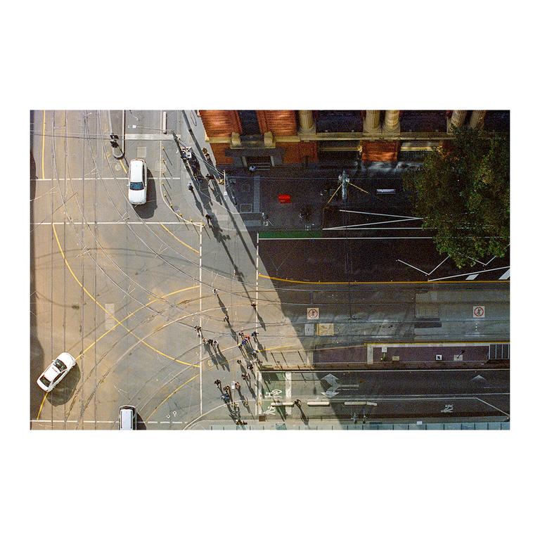 An aerial view shows a street with cars, people, and tram tracks next to a building and a tree.
