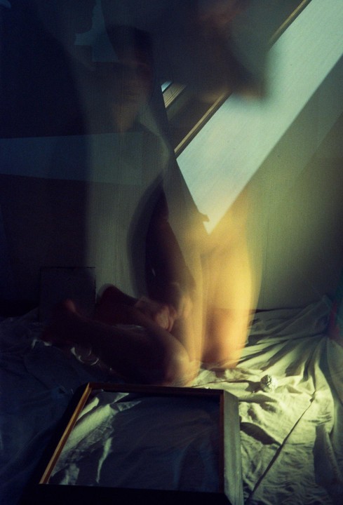 A blurry figure sits on a bed with a wooden frame in the foreground and light streaming in from the right.