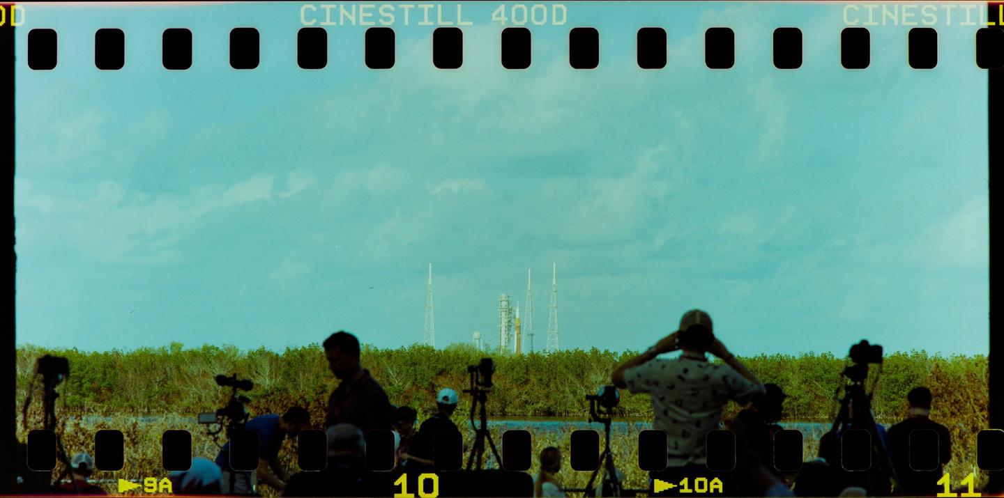 A group of people are gathered with cameras and tripods, looking towards a rocket and launch towers in the distance.