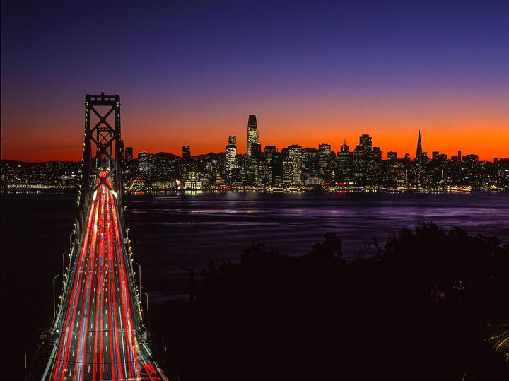A bridge with light trails from cars leads to a city skyline at dusk.