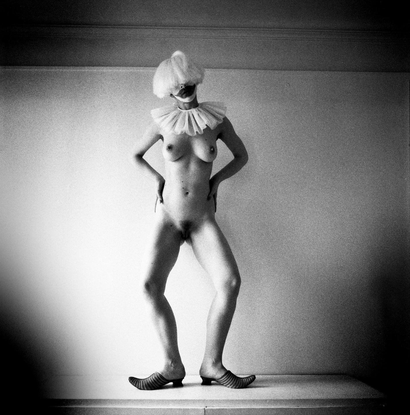 A nude woman with a clown wig, ruff, and striped shoes stands with her hands on her hips.