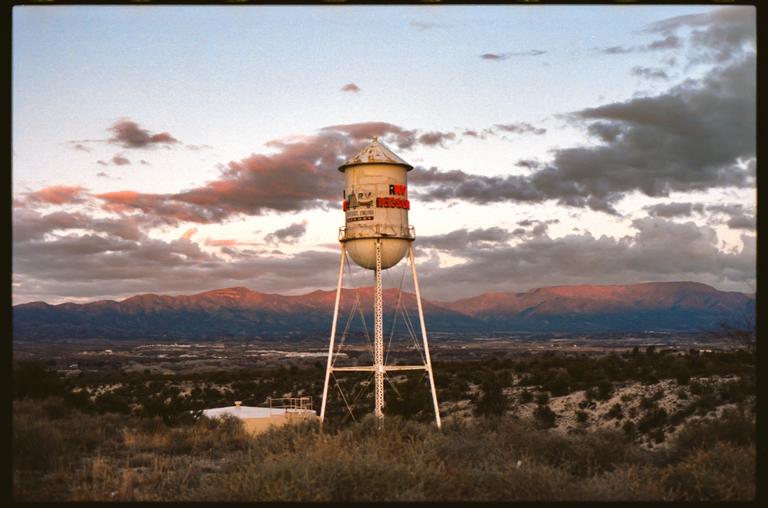 A water tower stands on a hill overlooking a valley and mountains under a cloudy sky at dusk.