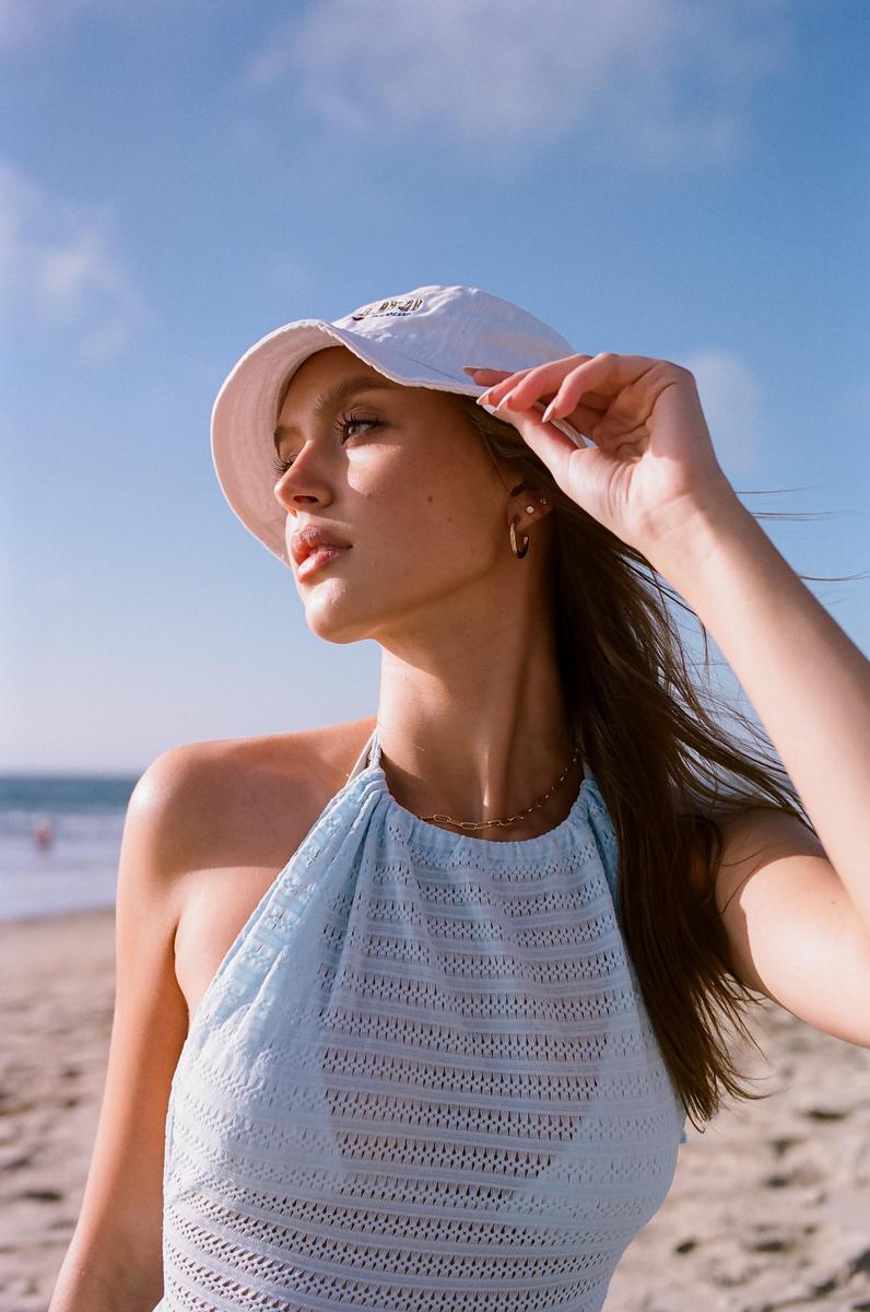 A woman wearing a bucket hat and a light blue top stands on a beach with the ocean in the background.