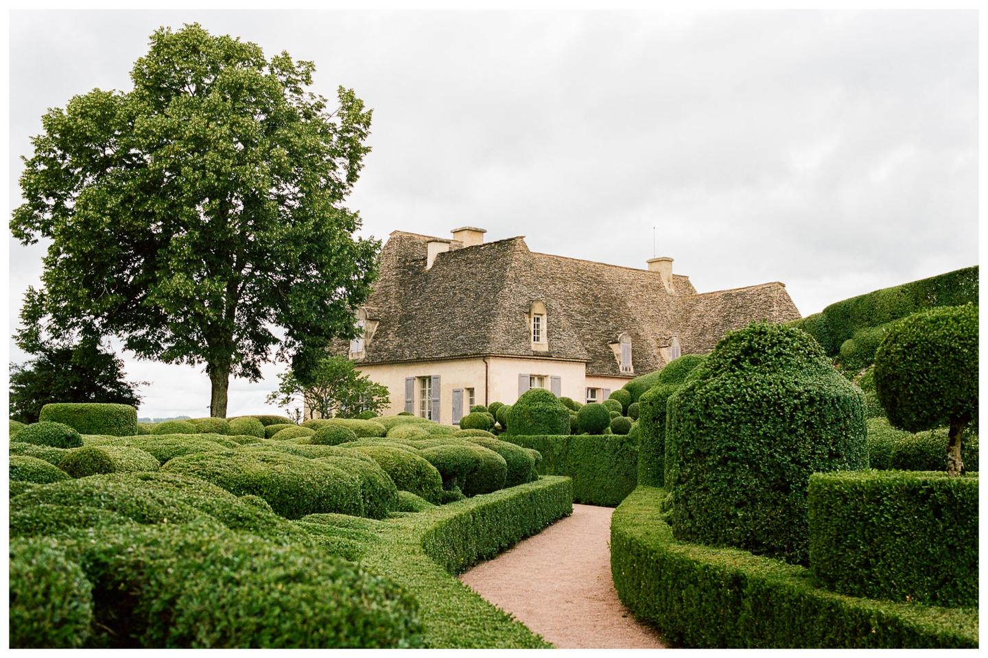 A large house with a stone roof is surrounded by an elaborate garden with sculpted hedges and a winding path.