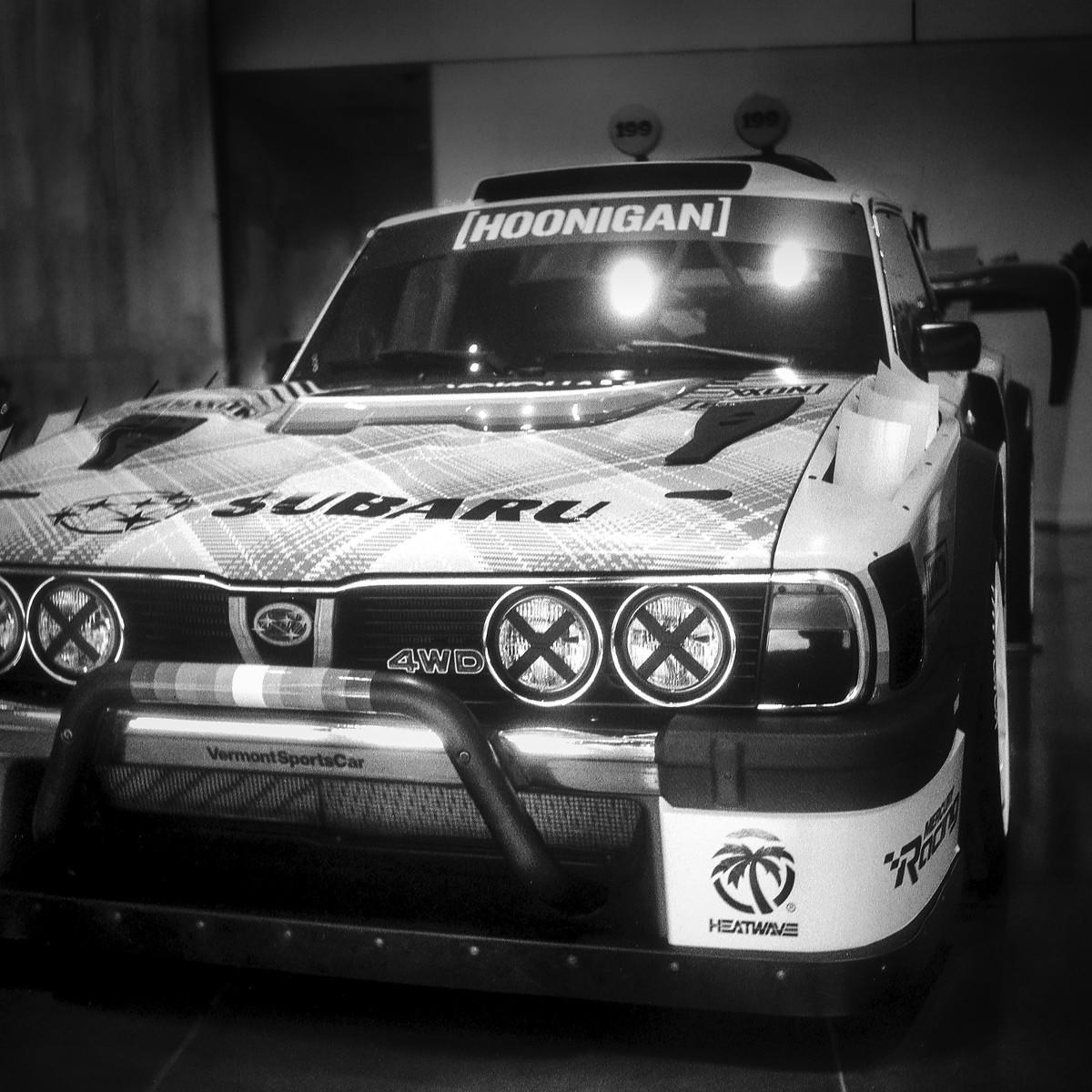 A black and white photograph shows the front of a Subaru rally car with a "Hoonigan" banner on the windshield.
