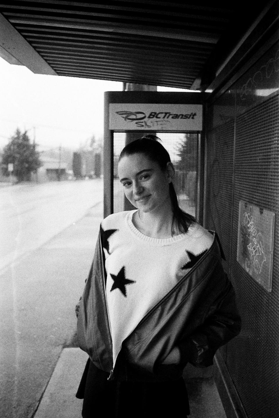 A woman wearing a star-patterned sweater and a jacket stands at a bus stop.