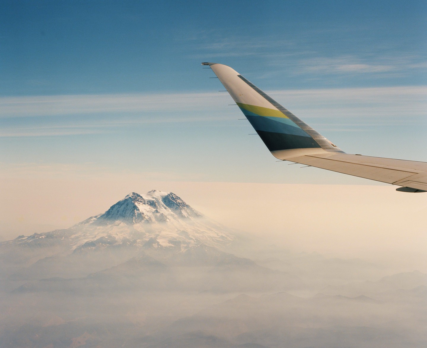 An airplane wing is visible above a snow-capped mountain peak shrouded in haze.