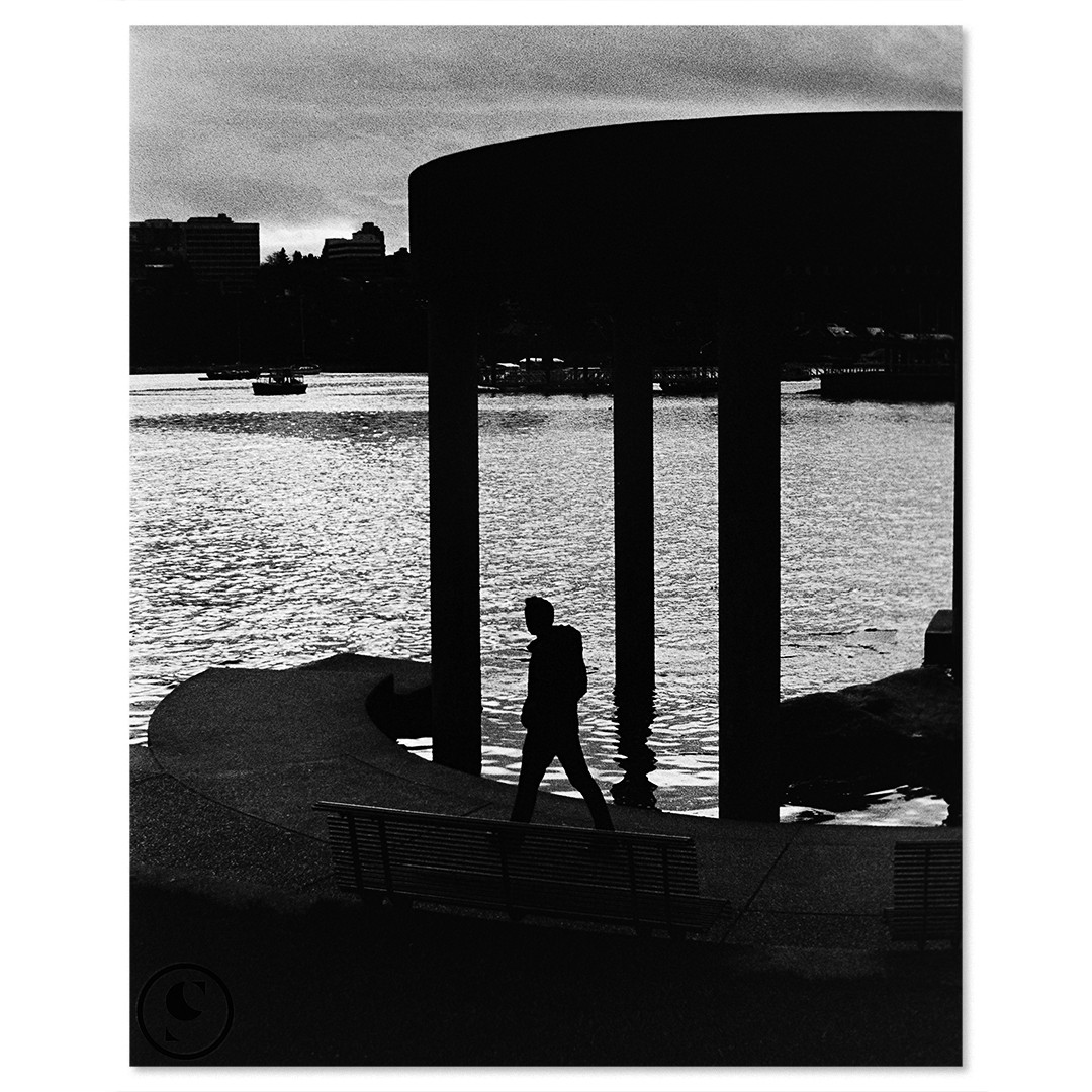A man walks along a waterfront path with a city skyline in the background.