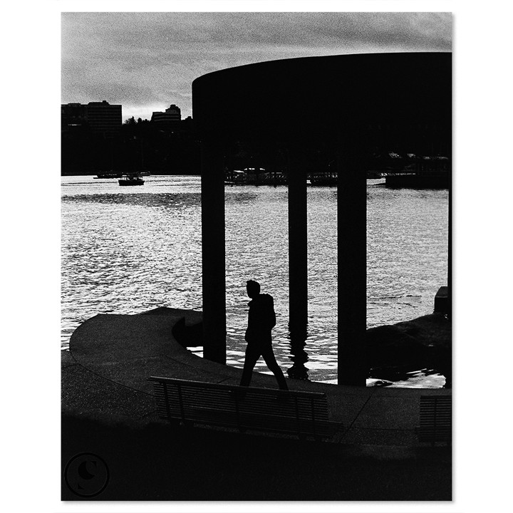 A man walks along a waterfront path with a city skyline in the background.