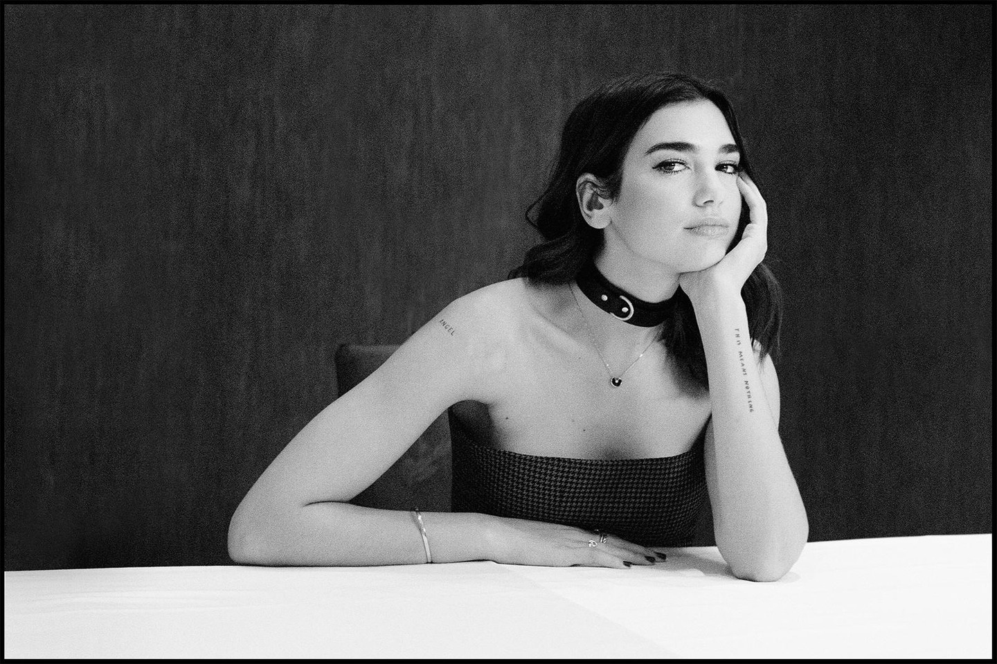 A woman with dark hair and a choker necklace rests her chin on her hand while sitting at a table.