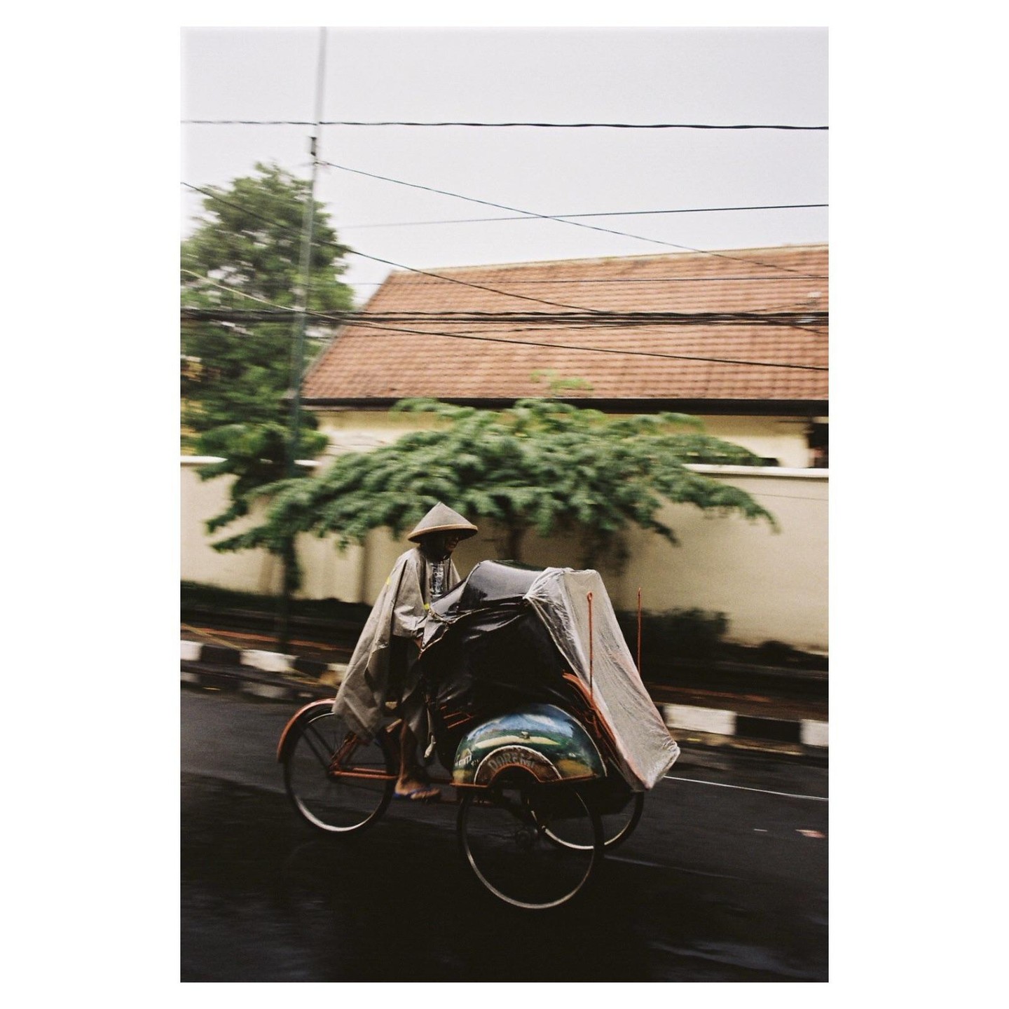 A man wearing a conical hat and poncho rides a rickshaw on a wet street.