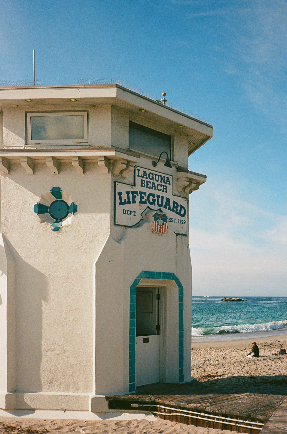 A white lifeguard tower with blue trim stands on a sandy beach next to the ocean under a clear sky.