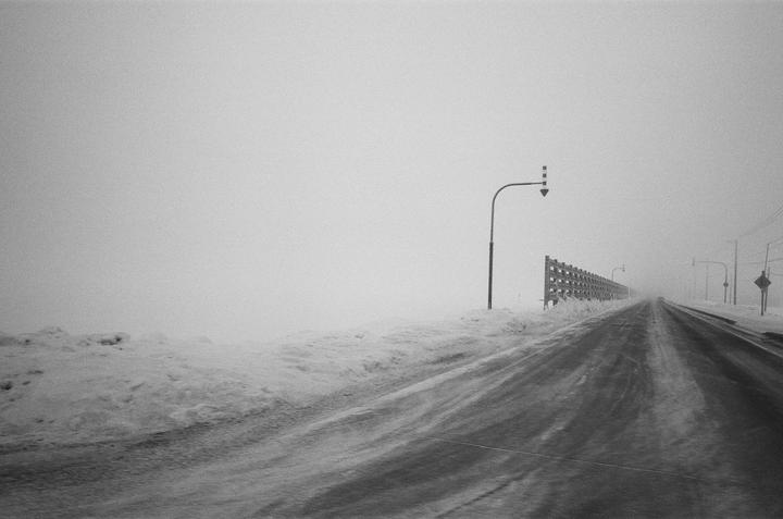 A snow-covered road stretches into the distance under a hazy sky, with a street light and a wooden fence visible on the right.