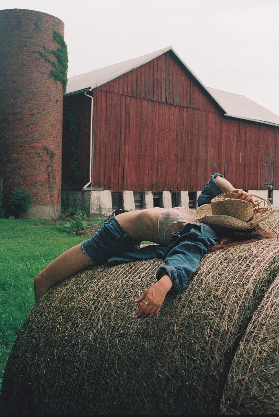 A woman wearing a cowboy hat, denim jacket, and shorts lies on a hay bale in front of a red barn and brick silo.