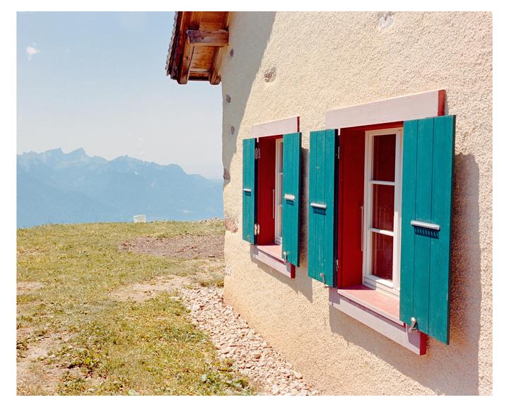 A building with red window frames and green shutters stands on a grassy hill overlooking mountains.