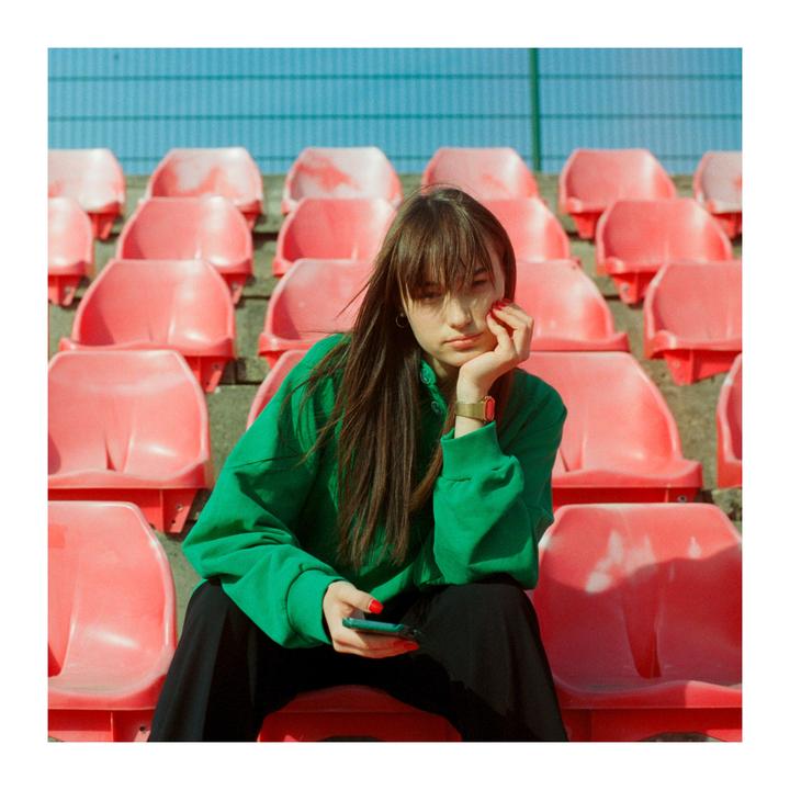 A woman in a green sweatshirt sits on a red stadium seat, holding a phone in her lap.
