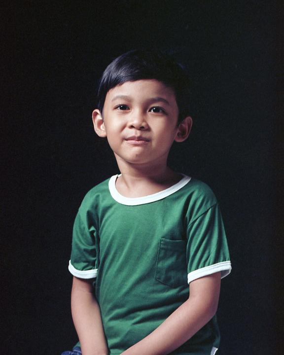 A young boy in a green shirt with white trim looks directly at the viewer.