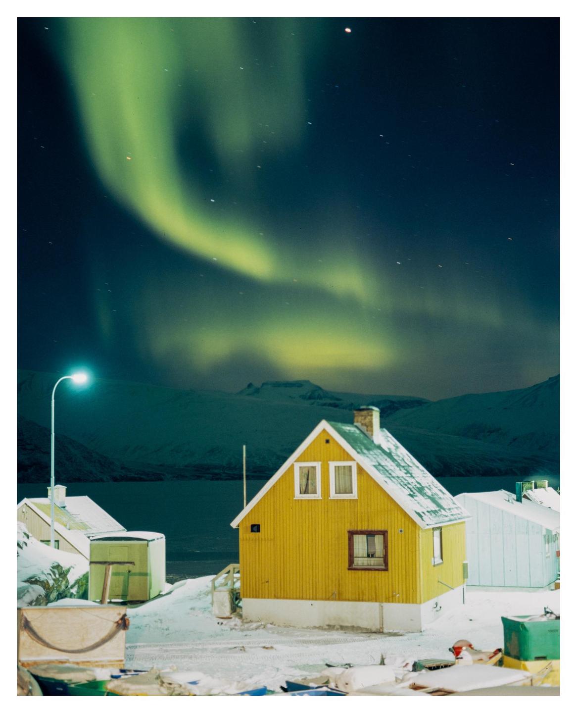 A yellow house with a snow-covered roof stands in a snowy landscape under a night sky with aurora borealis.