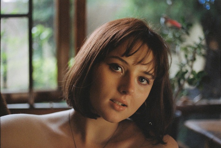 A woman with short hair looks at the viewer, with a window and greenery in the background.