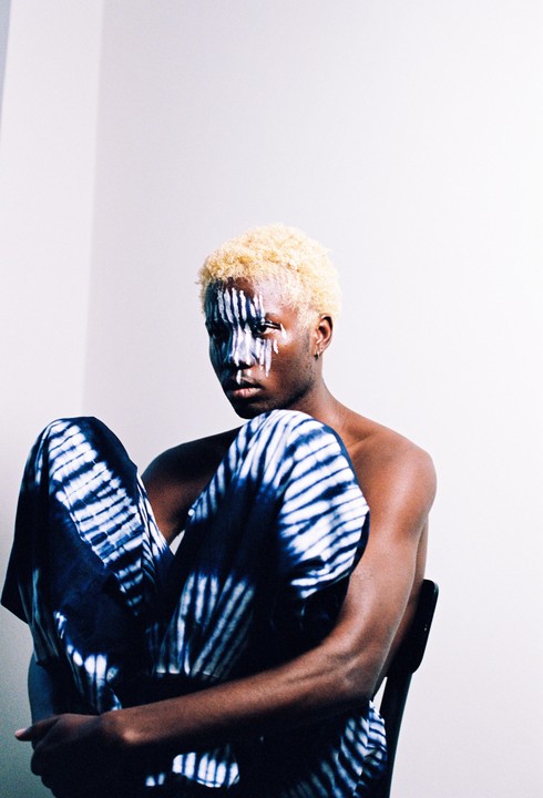 A man with bleached hair and white face paint sits on a chair, wearing blue and white patterned pants.