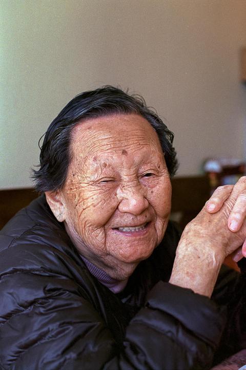 A smiling elderly woman with dark hair and a dark jacket is captured in a close-up portrait.