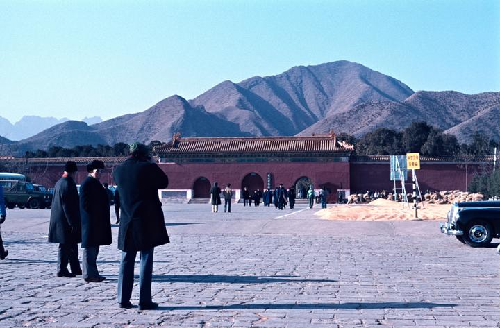 Several people stand on a paved area in front of a red building with a tiled roof, with mountains in the background.